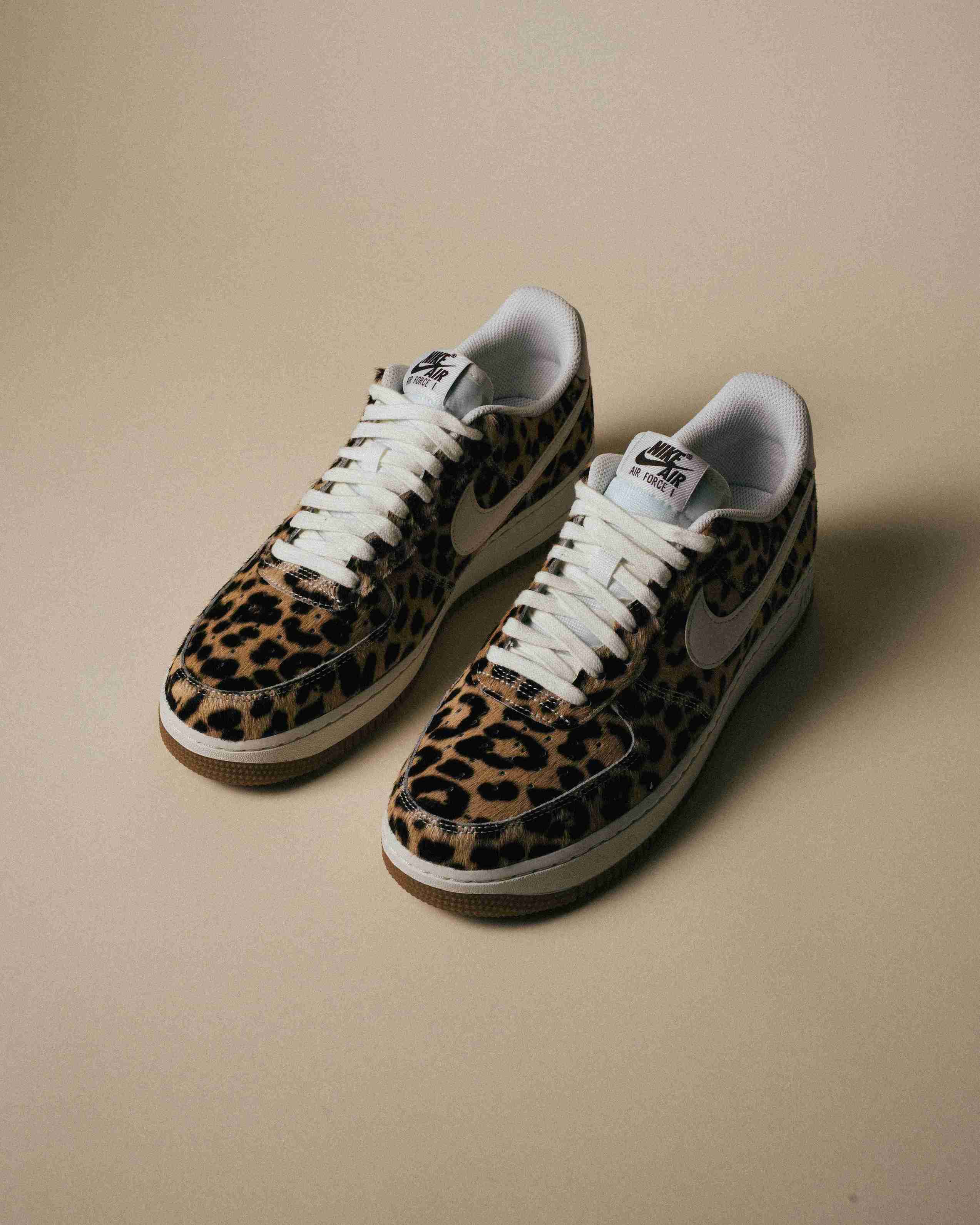 Paire de sneakers Nike Air Force 1 à motif léopard, placée à côté d’une boîte Nike grise sur un fond beige, vue artistique avec lumière douce.